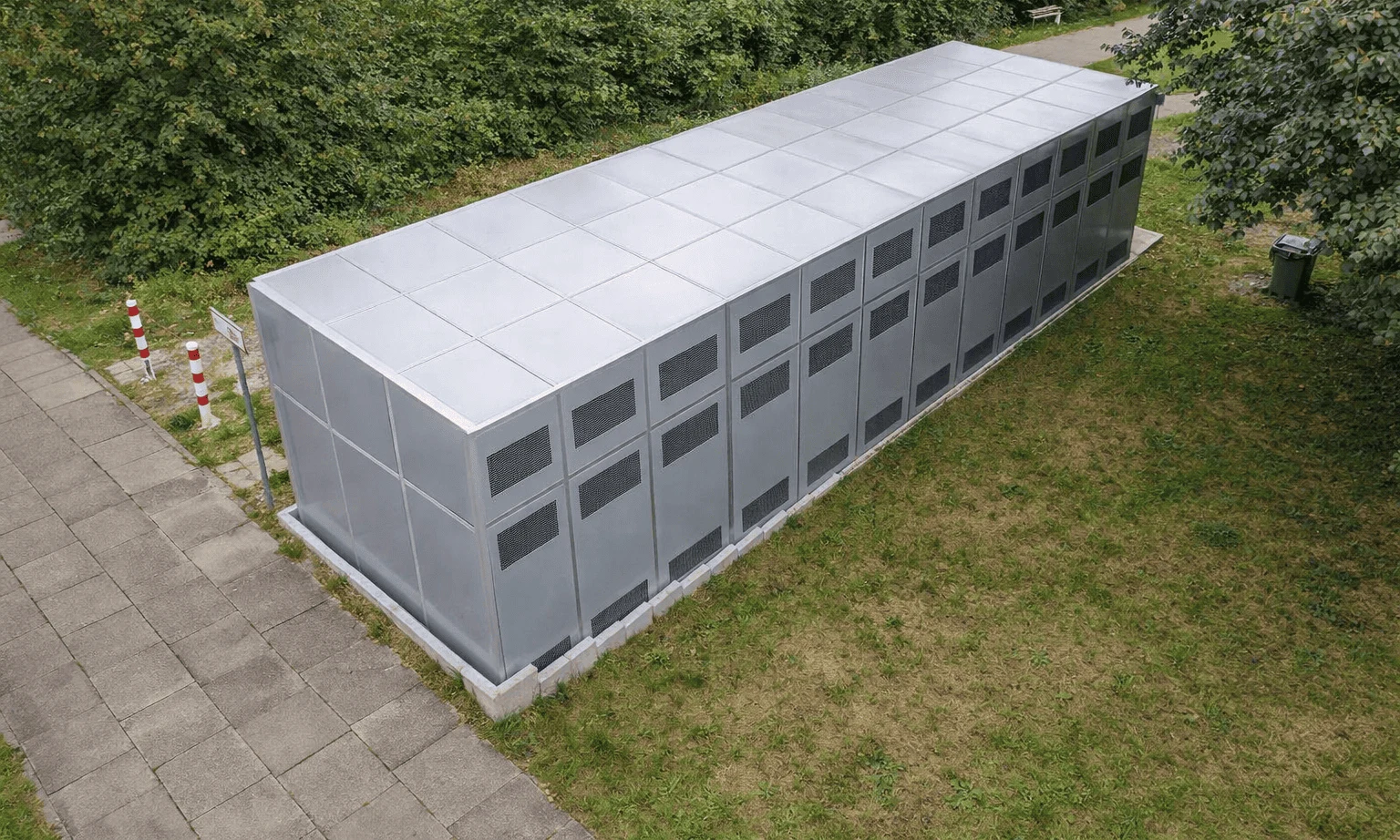 Durchgehende 12 Meter lange Schallhaube aus Aluminium-Paneelen mit Lüftungsgittern für vier Weishaupt-Wärmepumpen, auf einer Grünfläche neben einem Gehweg